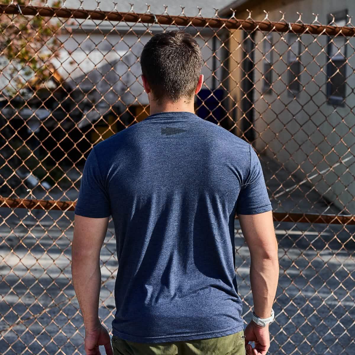 Man in navy GORUCK t-shirt standing outdoors by a rusted chain-link fence
