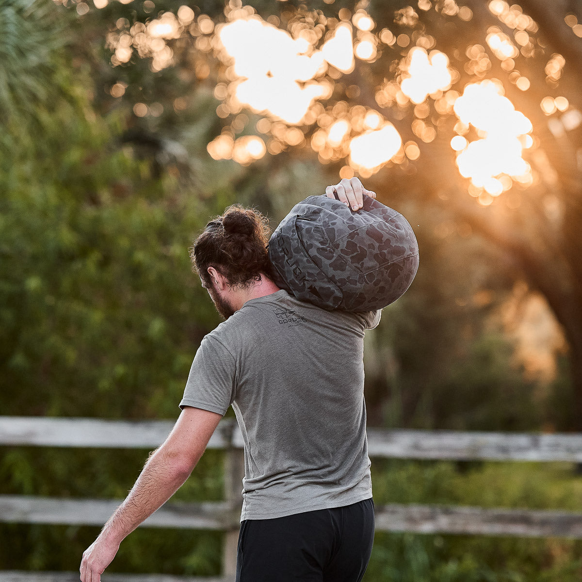 A man with a bun carries a Sand Medicine Ball on his shoulder outdoors at sunset, standing near a wooden fence.