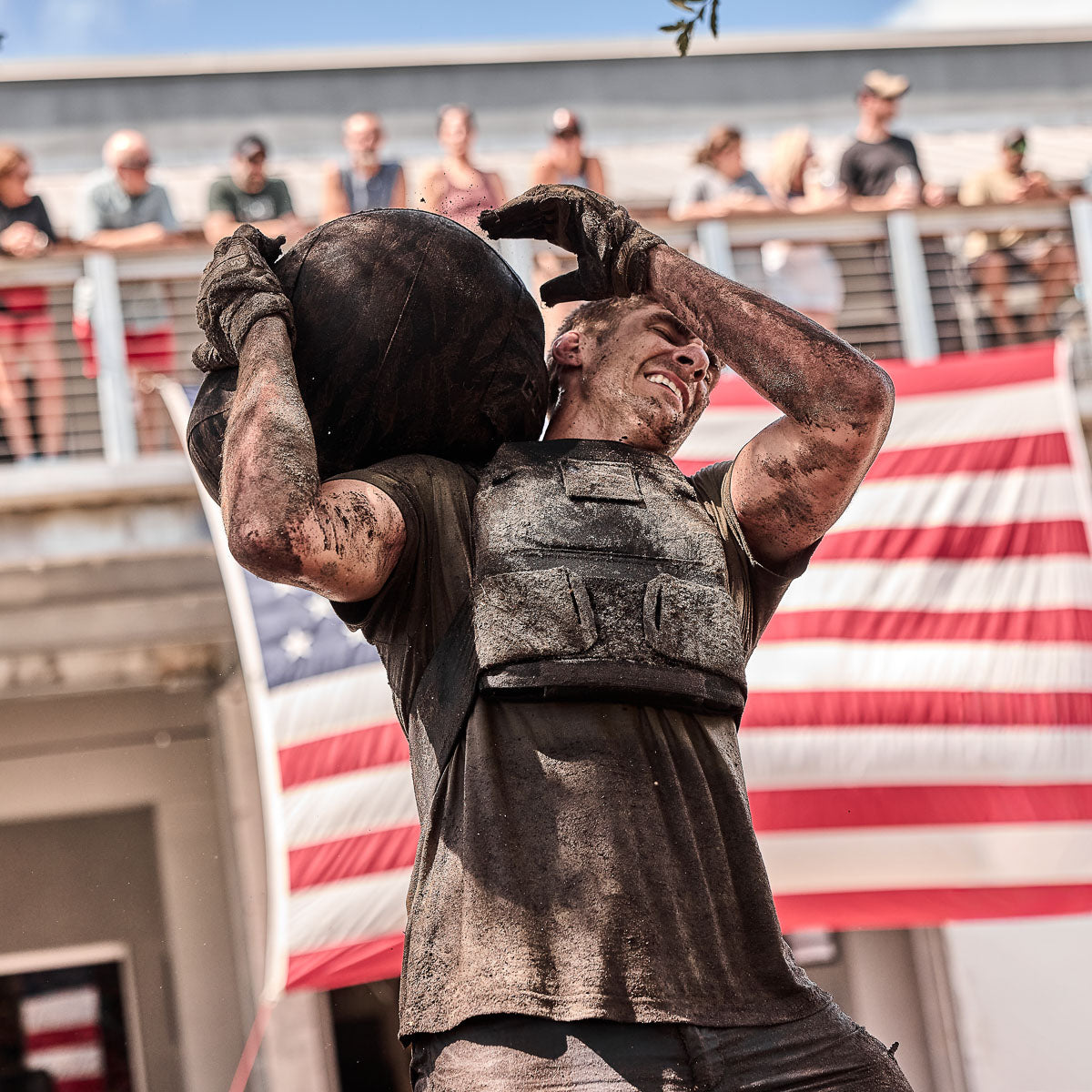 A man in a weighted vest lifts a heavy ball, covered in dirt, with a large American flag behind him.