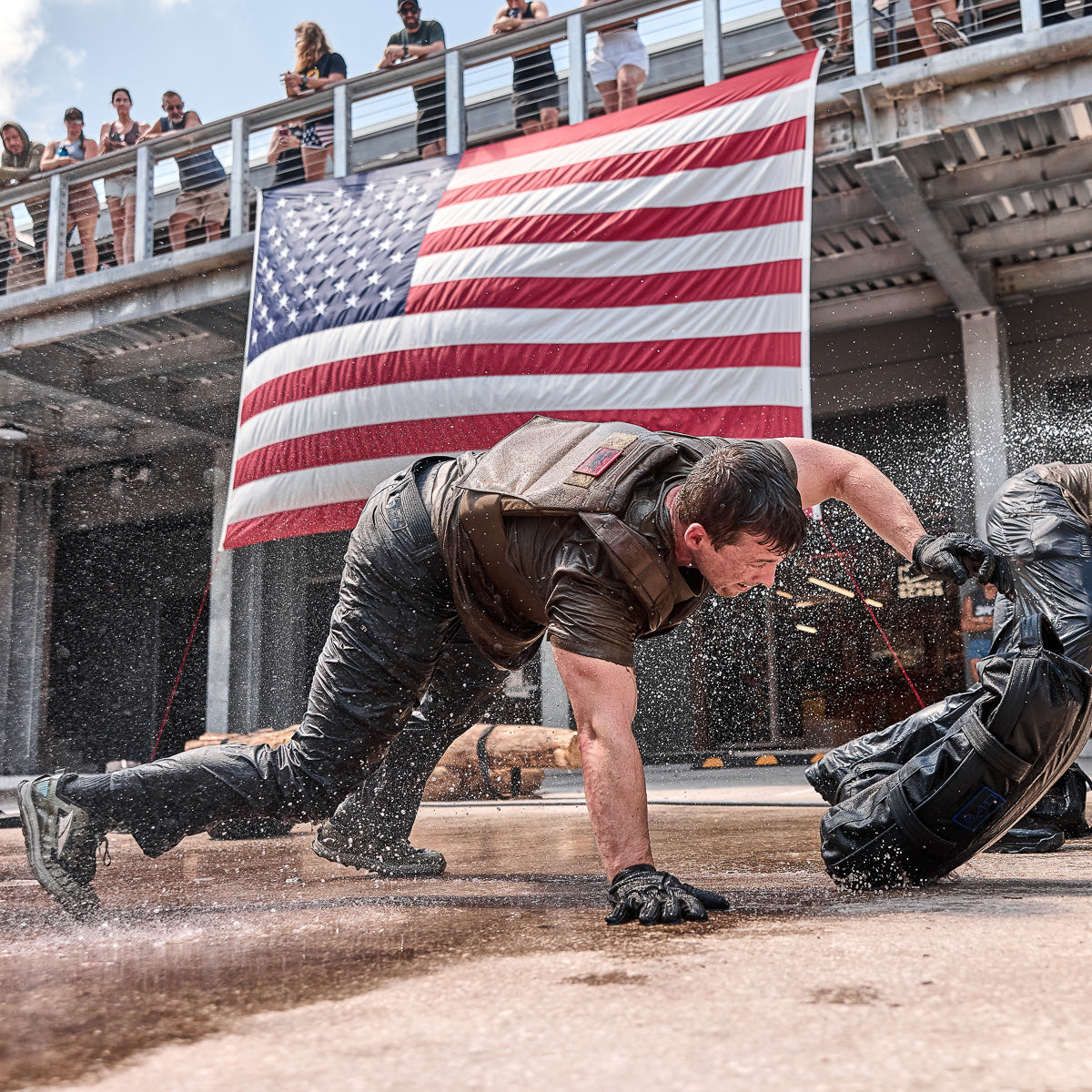 Man in wet clothes crawling on the ground, American flag and spectators in the background.