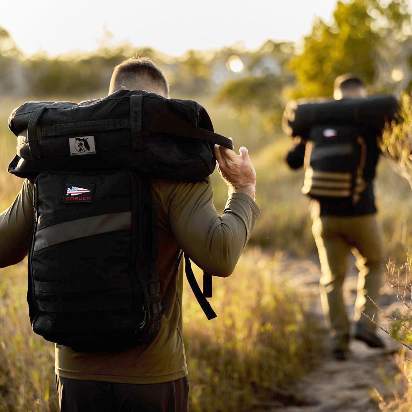 Two people hike on a dirt path carrying large black backpacks in a sunlit outdoor setting.