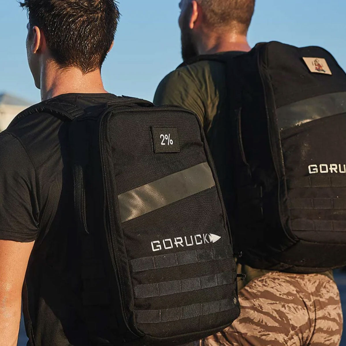 Two people wearing black GORUCK backpacks are walking outdoors in sunlight, seen from behind.