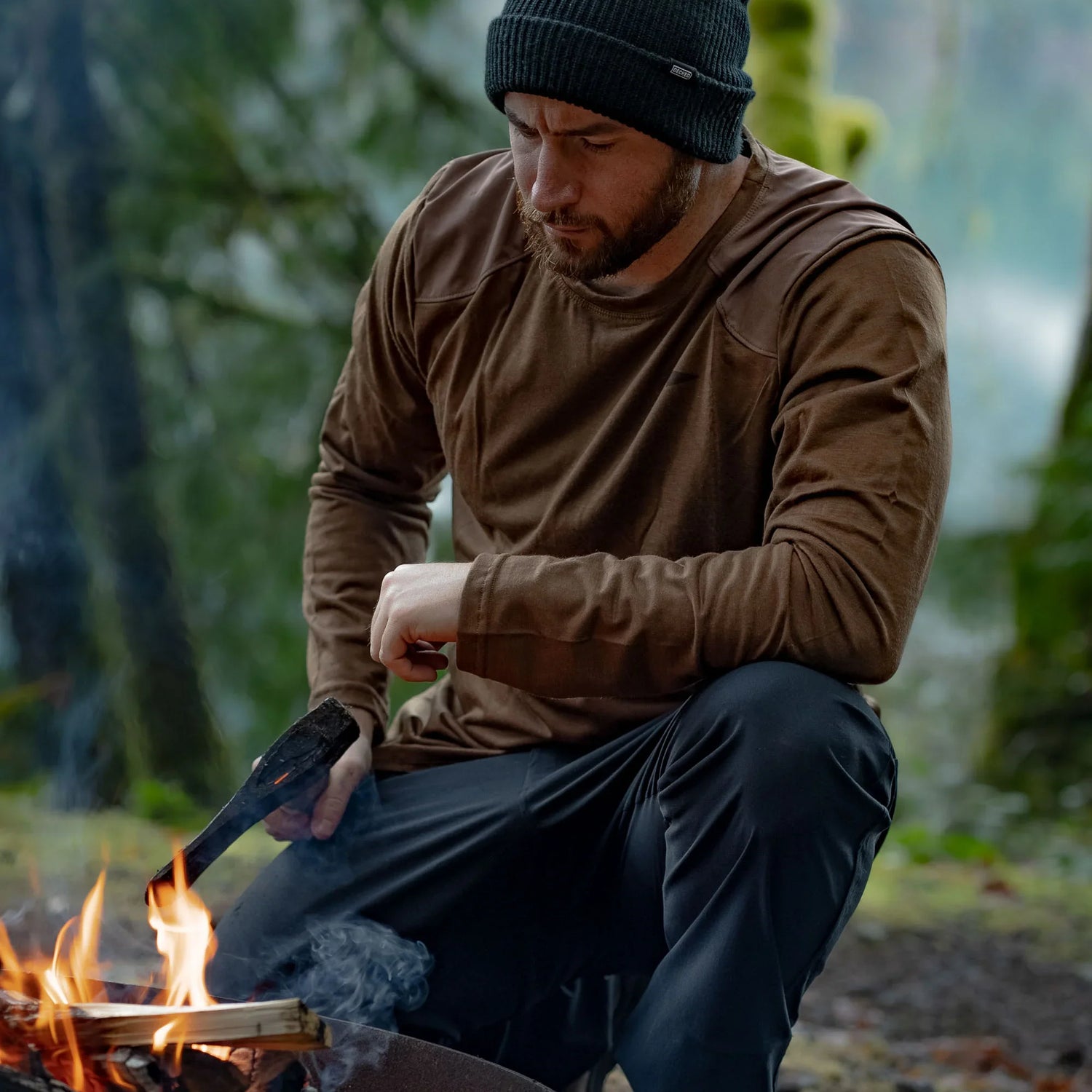 Man in GORUCK outdoor gear tending campfire in forest, wearing brown shirt and black beanie.
