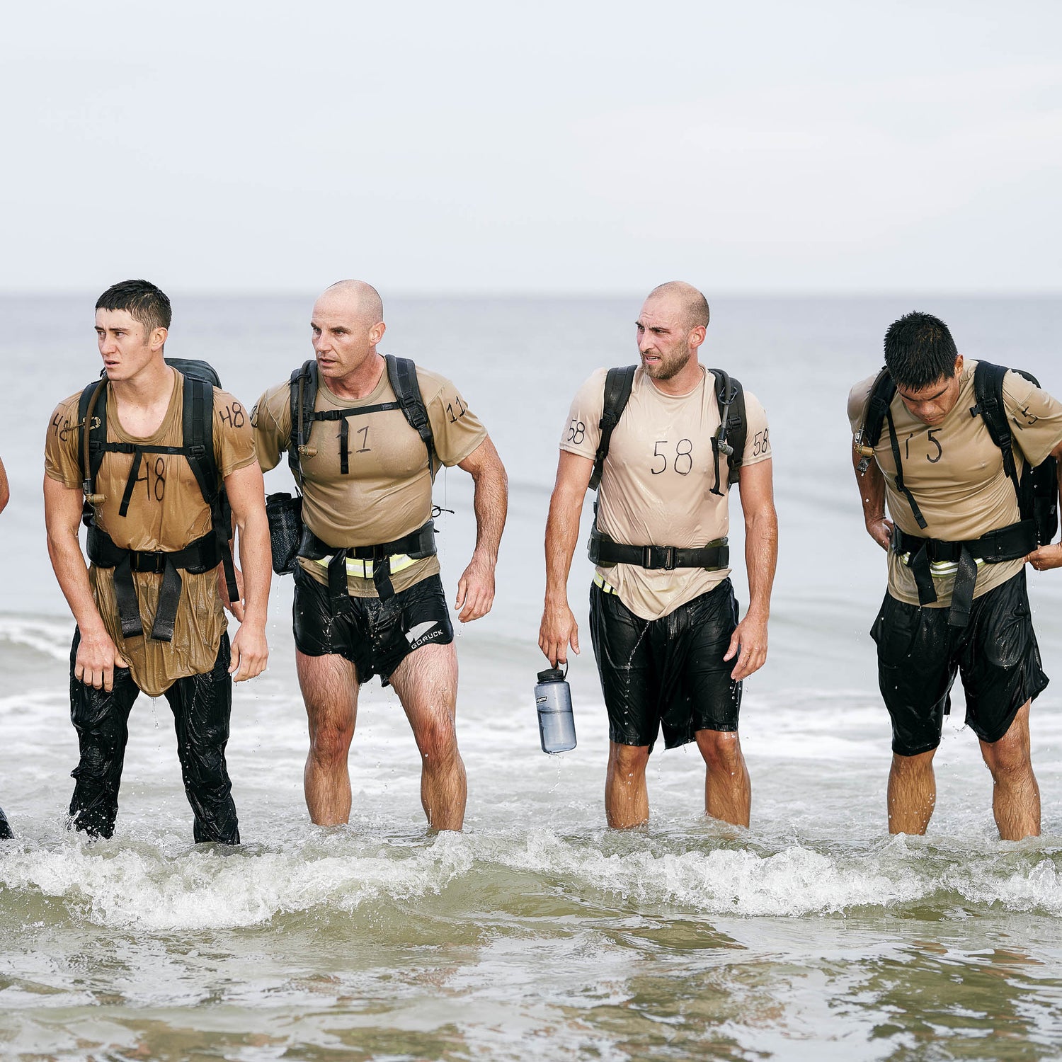Four men in wet athletic gear, each wearing a numbered shirt and a Padded Hip Belt backpack, stand in shallow ocean water, ready for serious rucking.