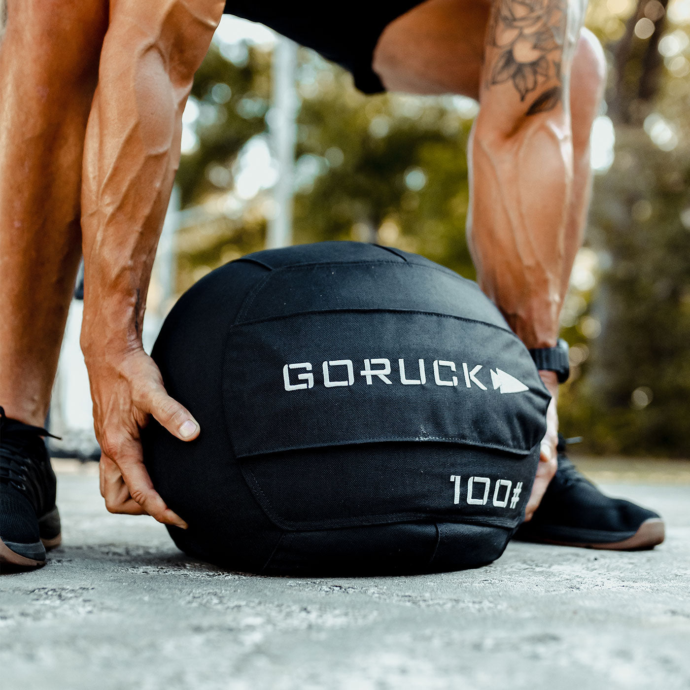 A person lifts a black GORUCK 100 lb sandbag outdoors, showing muscular arms and veins.
