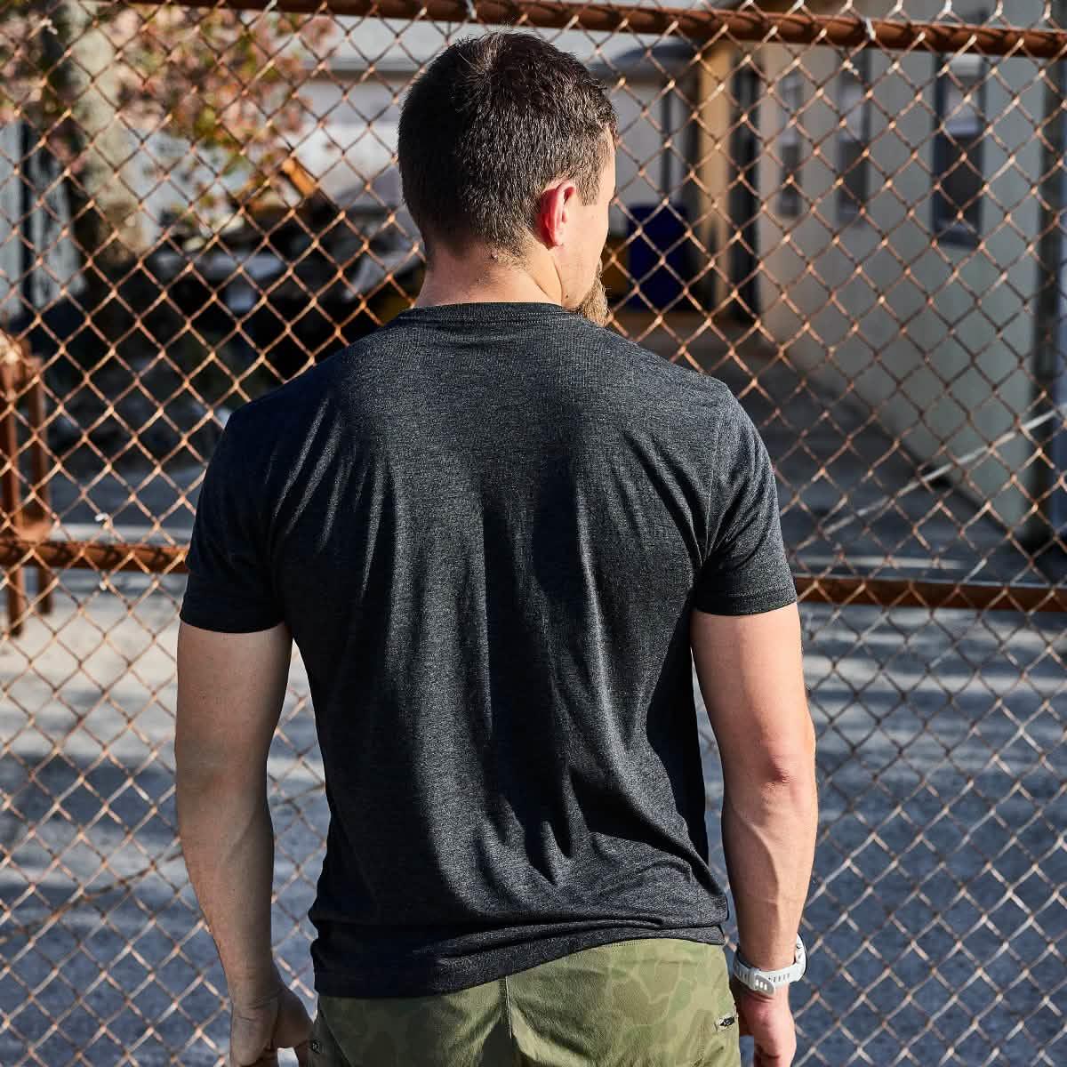 Man wearing GORUCK black athletic tee and camo shorts stands by chain link fence outdoors