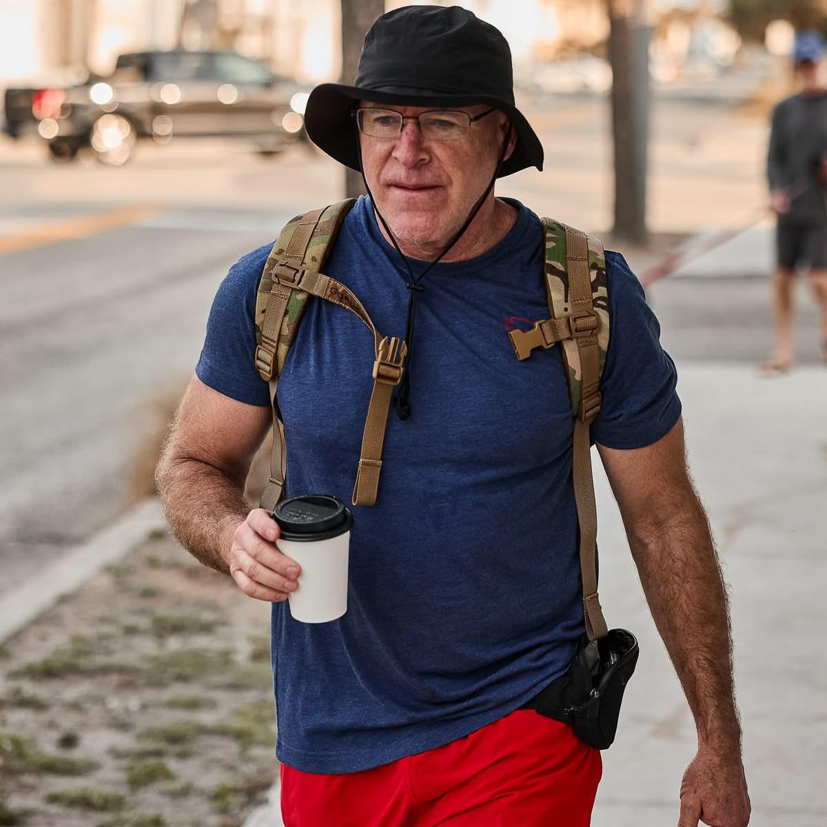 Man rucking in GORUCK navy Subtle Spearhead tee, tan backpack, carrying coffee outdoors