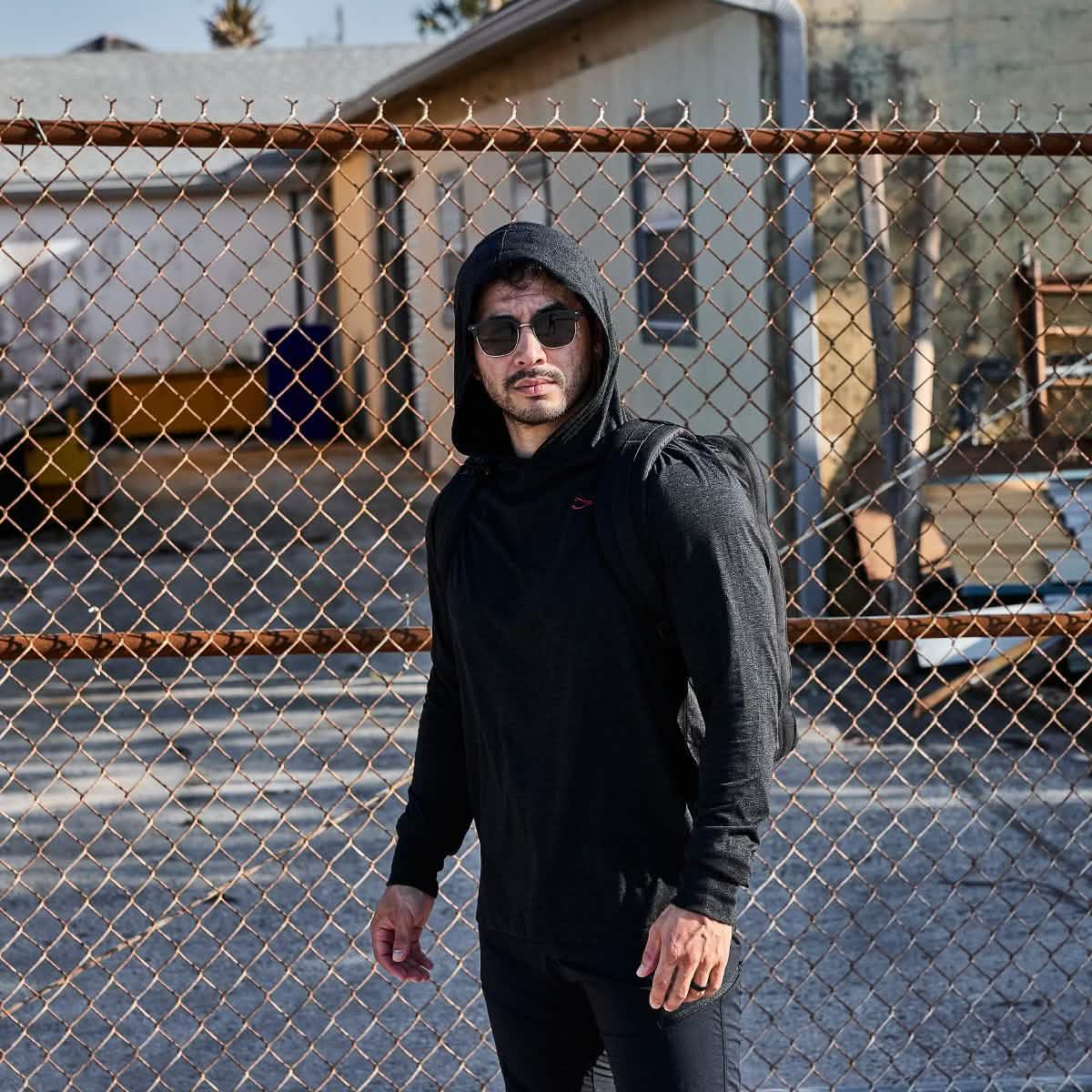 Man wearing black GORUCK hoodie and backpack, standing outdoors by a chain link fence