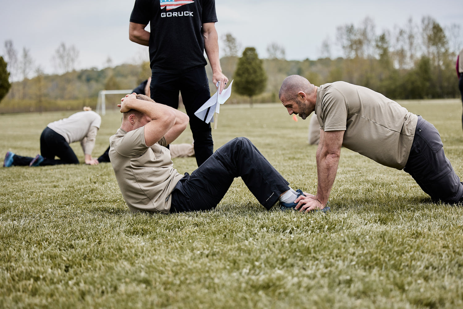 Two men exercise outdoors; one does sit-ups while the other holds his feet on a grassy field.