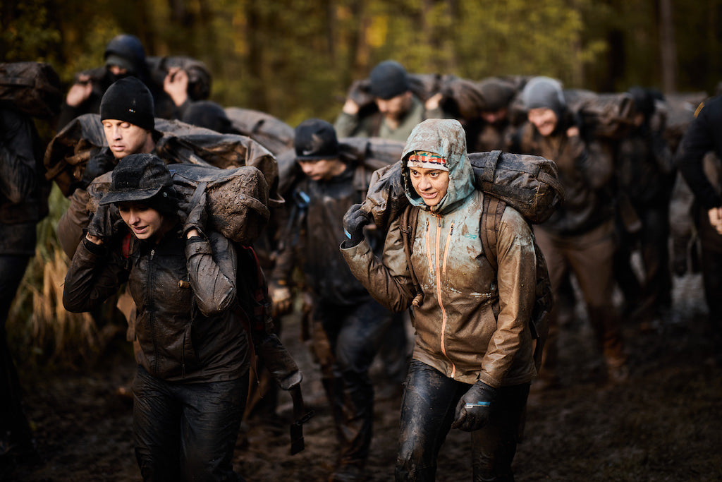 A group of people in muddy clothes carry heavy sacks on their shoulders through a forested area.