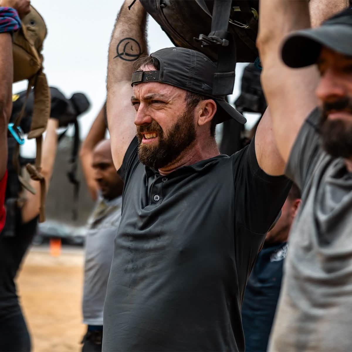 Group of men in GORUCK hats and gear lifting rucksacks overhead during outdoor rucking event