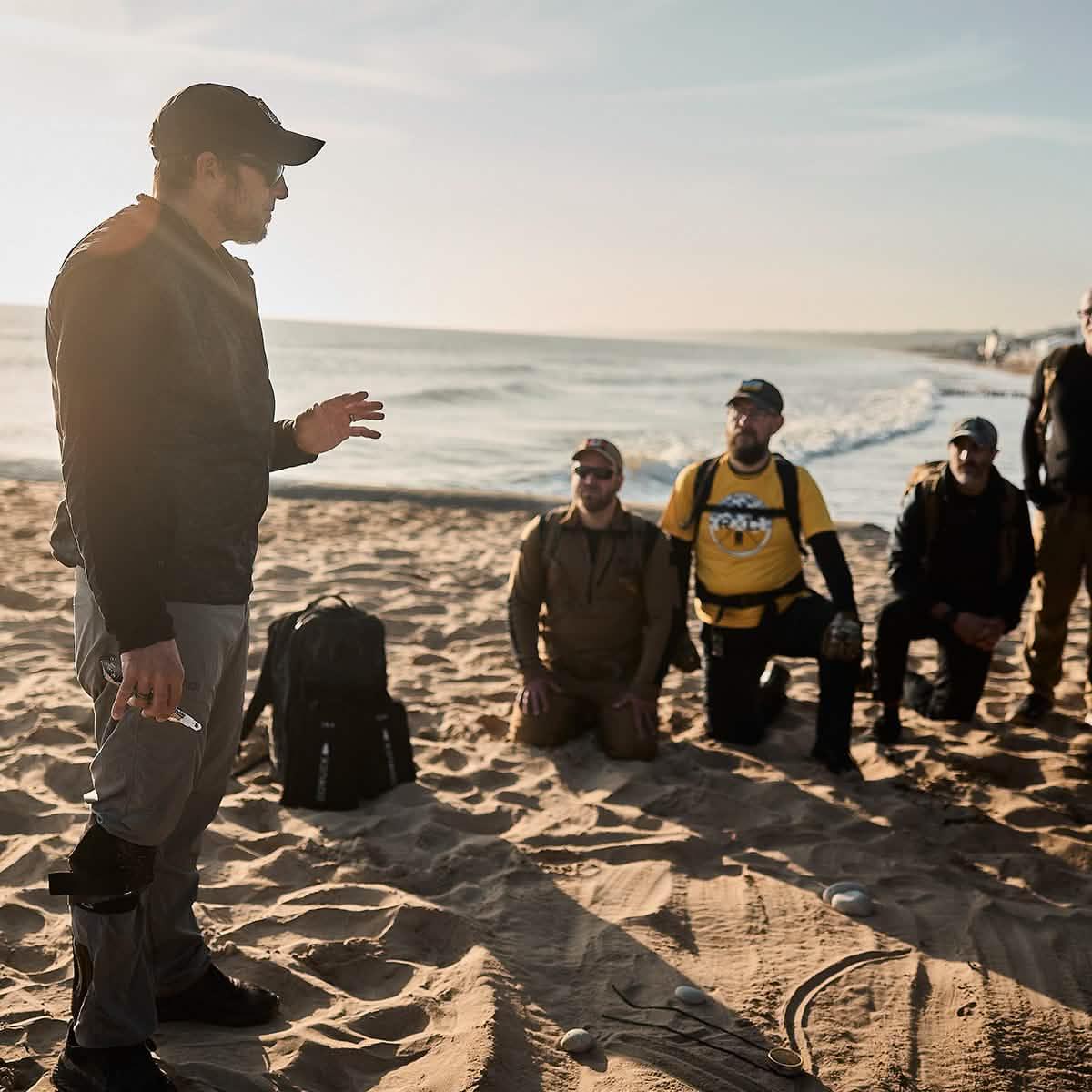 GORUCK rucking group training on sandy beach near ocean, wearing outdoor tactical gear