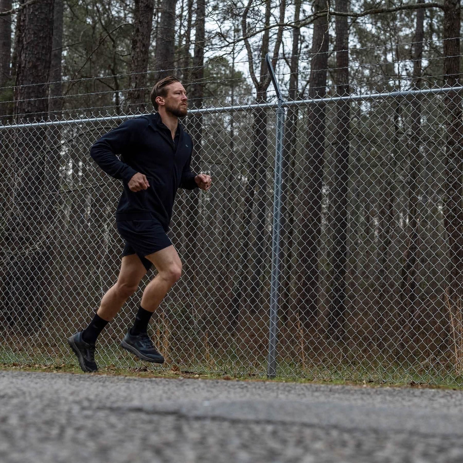 Man running outdoors wearing GORUCK gear by a chain-link fence and pine trees