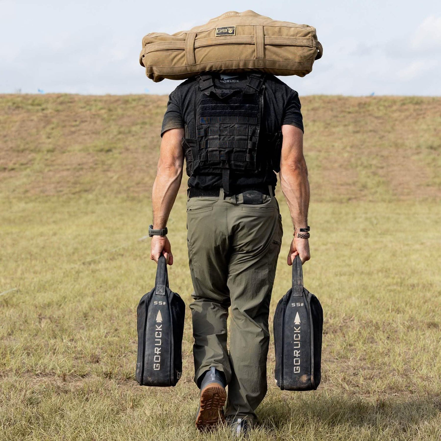 Man wearing a tactical vest carries GORUCK sandbags and gear outdoors on grass field