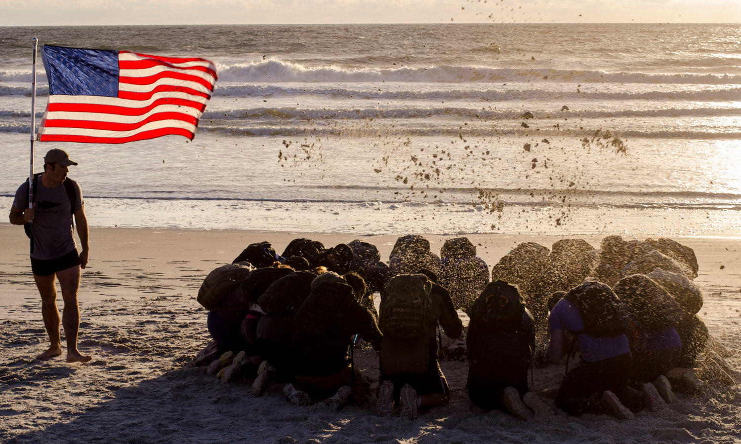 People kneel on the beach facing the ocean, while one person stands holding an American flag.