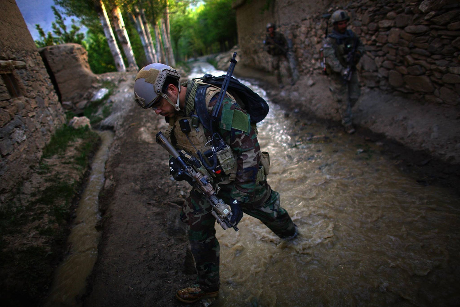 Three soldiers in camouflage gear walk through a muddy, narrow water path between stone walls and trees.