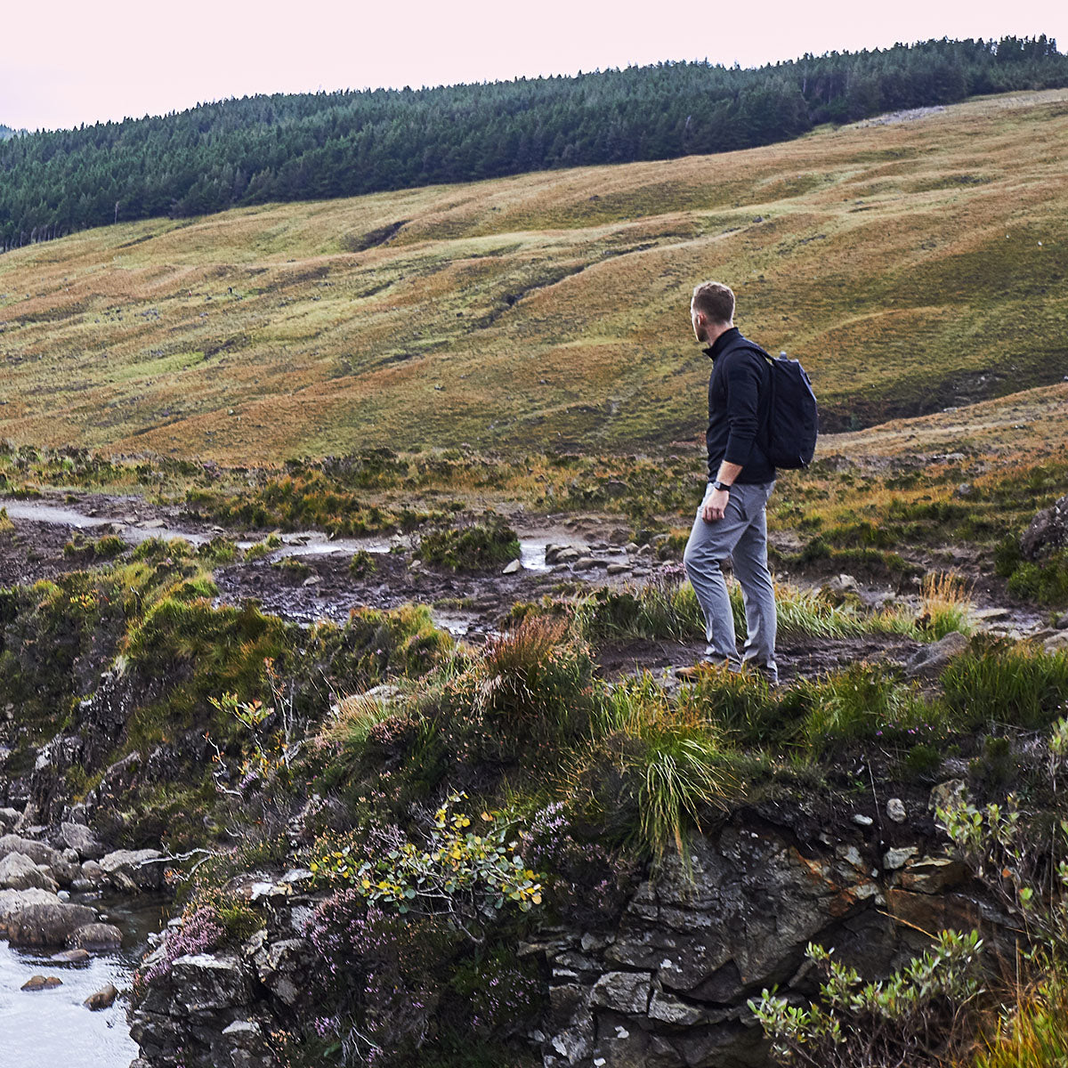 A man with a backpack stands on a grassy hill, looking out over a wide, rugged landscape.