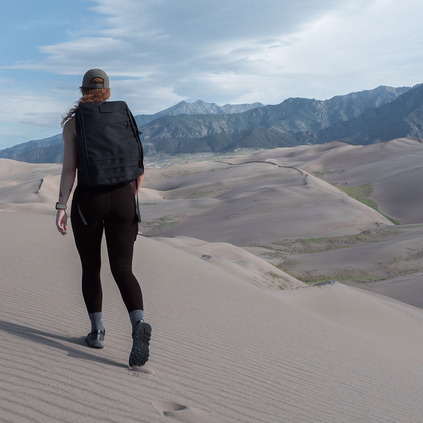 Person with a backpack hiking on sand dunes with mountains and cloudy sky in the background.