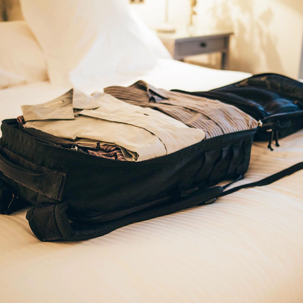 An open suitcase filled with neatly folded clothes sits on a white bed in a softly lit room.