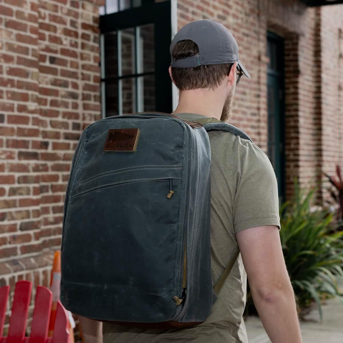 Man wearing GORUCK heritage slate rucksack outdoors, brick wall and plants in background