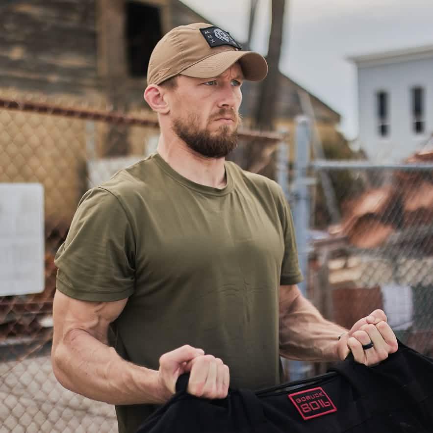 Man outdoors wearing a GORUCK hat and olive shirt, lifting rucking gear, rugged urban background