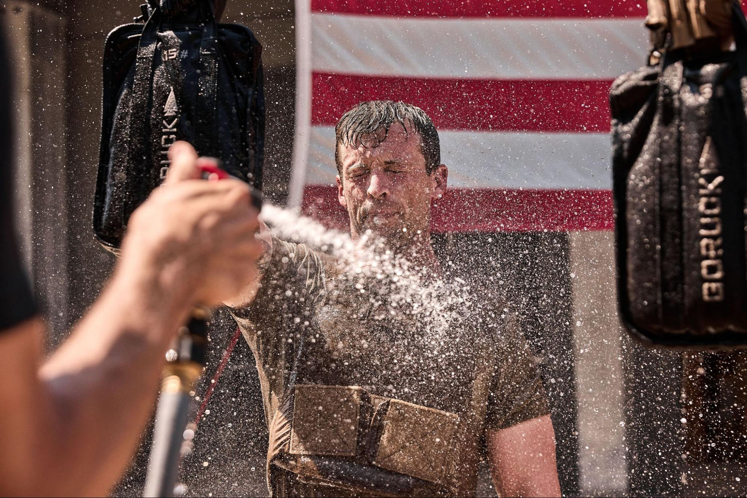 Man in wet clothes getting sprayed with water, with an American flag in the background.