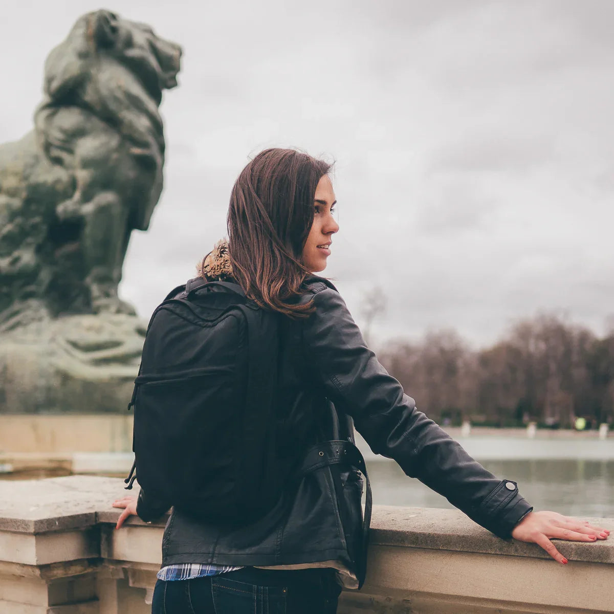 Woman wearing GORUCK backpack outdoors by a stone railing and lion statue, cloudy sky