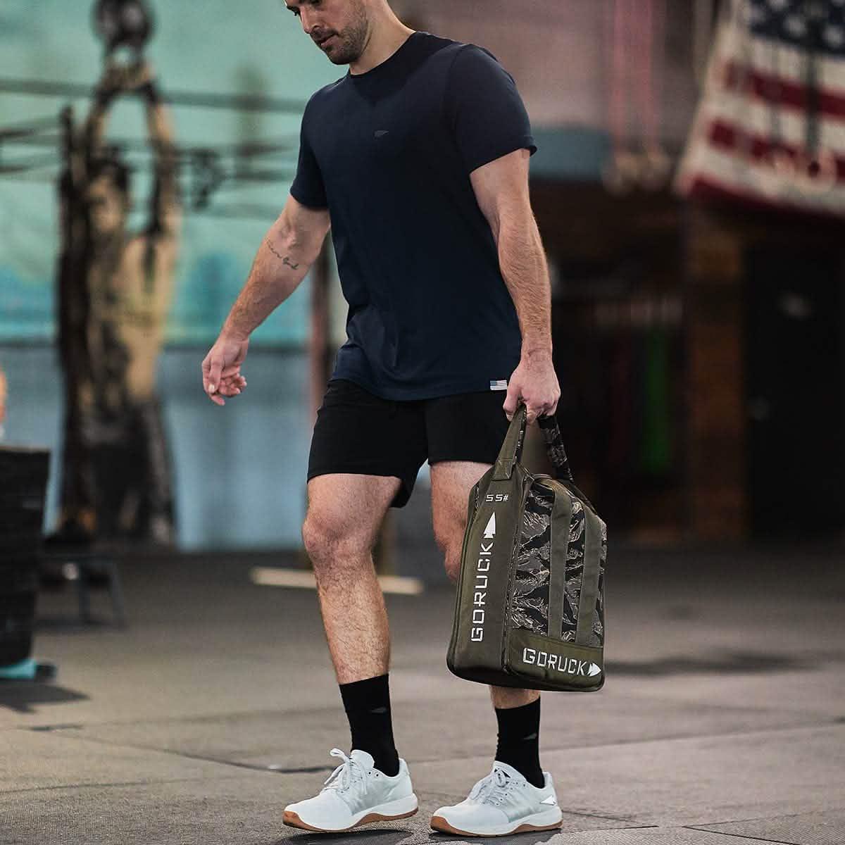 Man carrying GORUCK rucking gear bag in gym, wearing athletic shorts and t-shirt