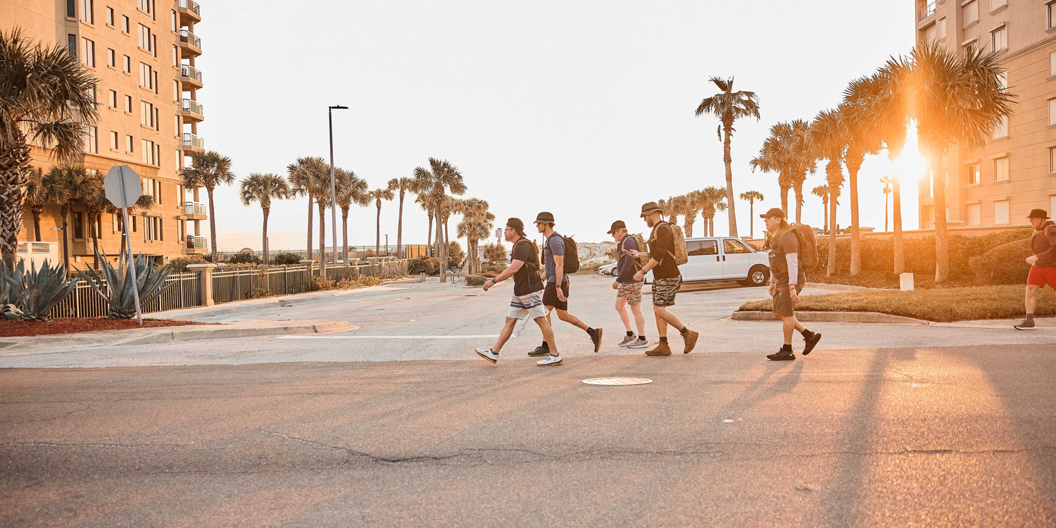 Six people walk across a street near palm trees and buildings at sunset.