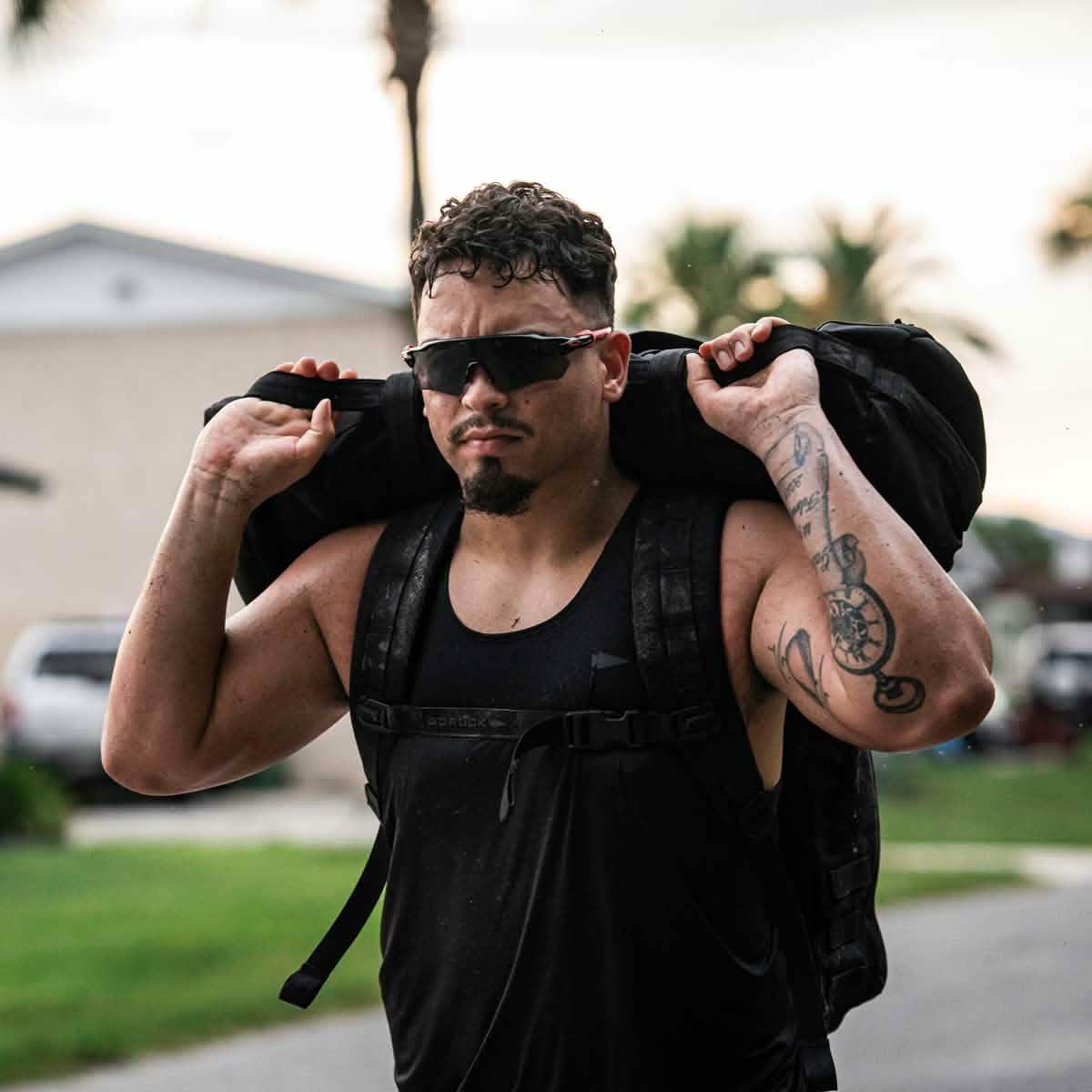 Man in black GORUCK performance tank carrying rucksack outdoors, wearing sunglasses