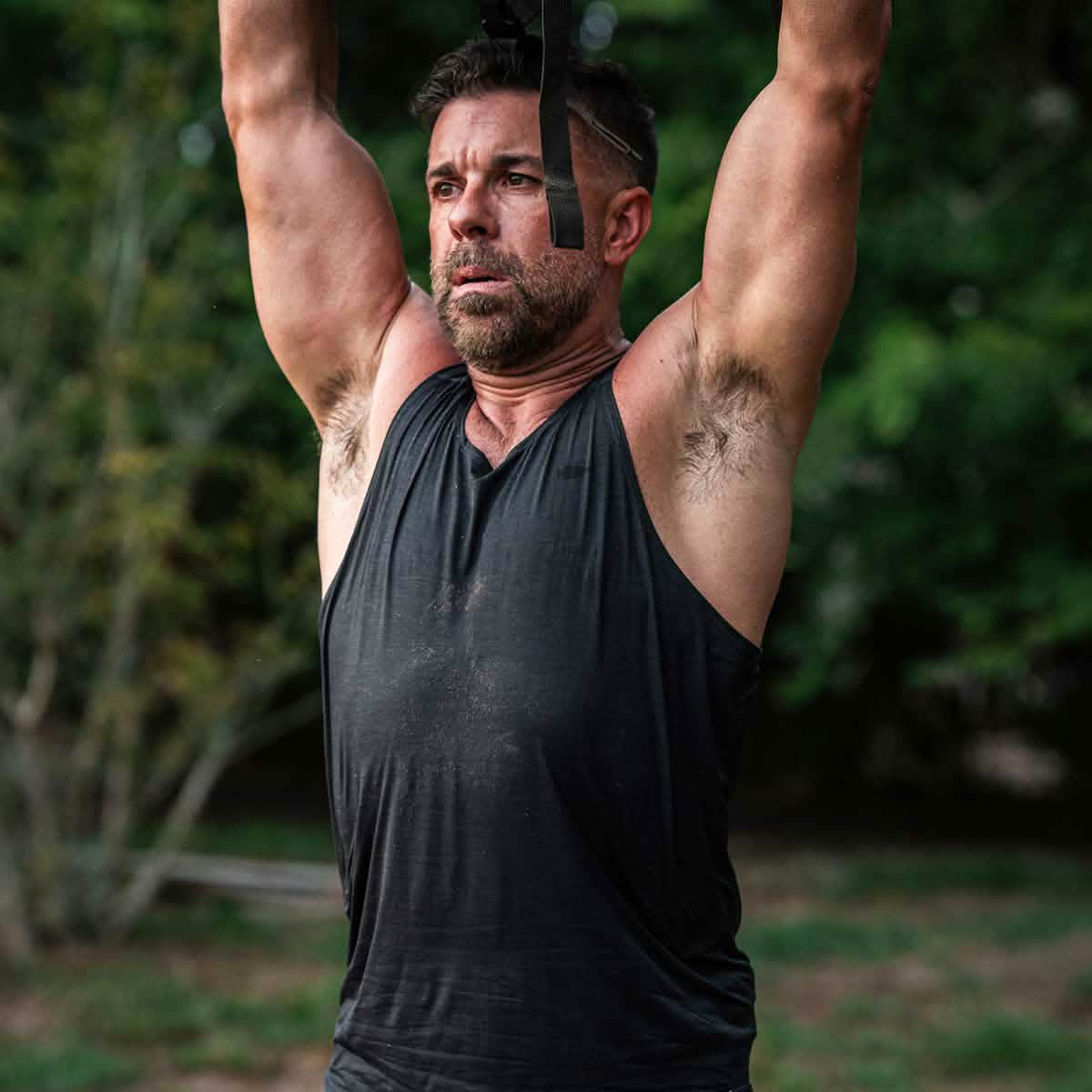 A man in a Men’s Performance Tank - ToughMesh works out outdoors, gripping overhead straps, with trees behind him.