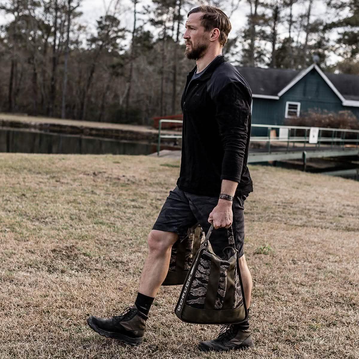 Man rucking outdoors with GORUCK gear, carrying weighted sandbags near a lake and cabin