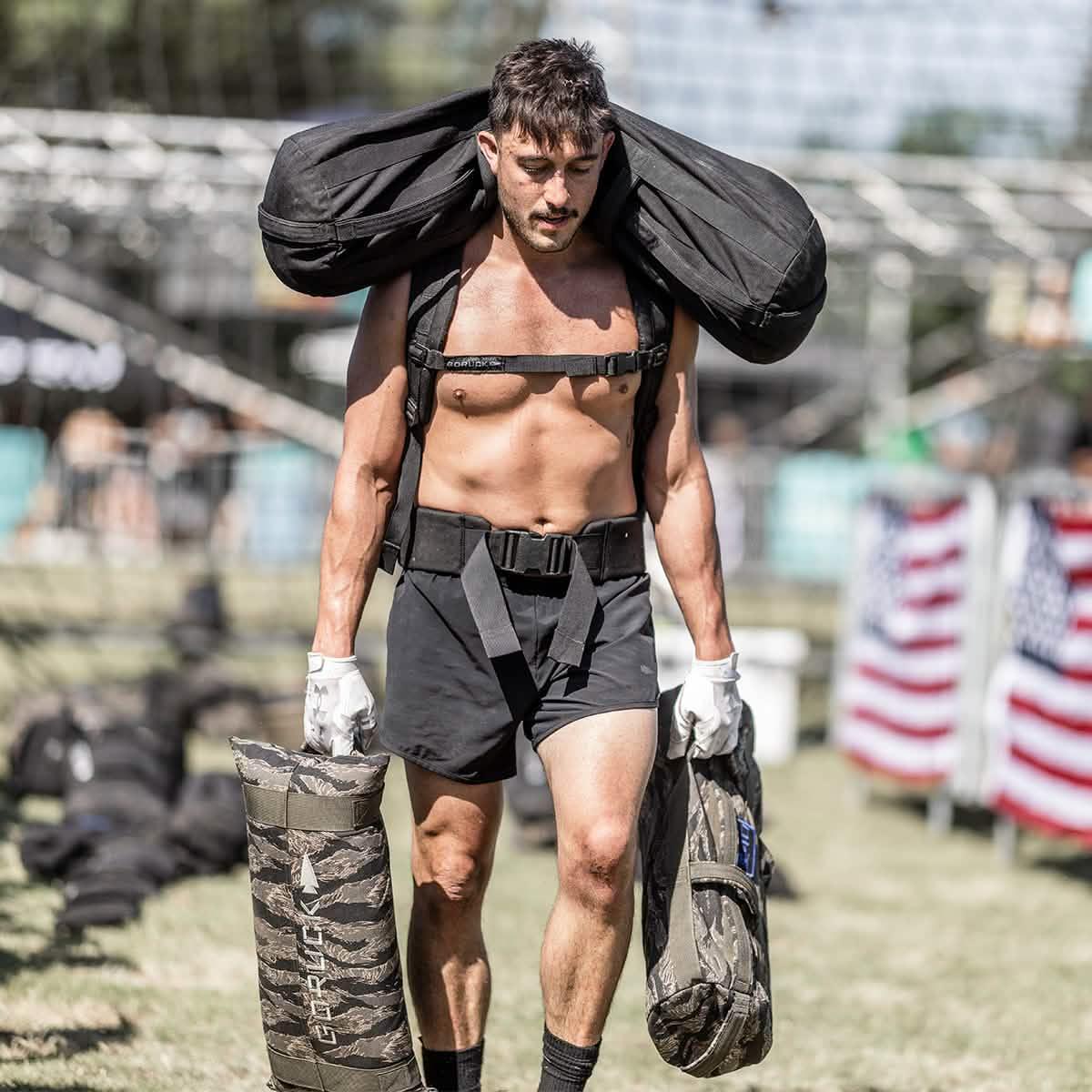 Man wearing black GORUCK shorts carrying sandbags at outdoor rucking event with USA flags