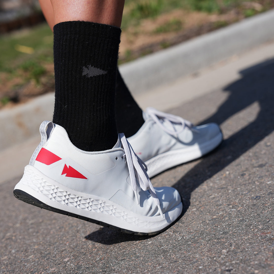 Close-up of a person wearing white running shoes with red accents and black socks, standing on a pavement.