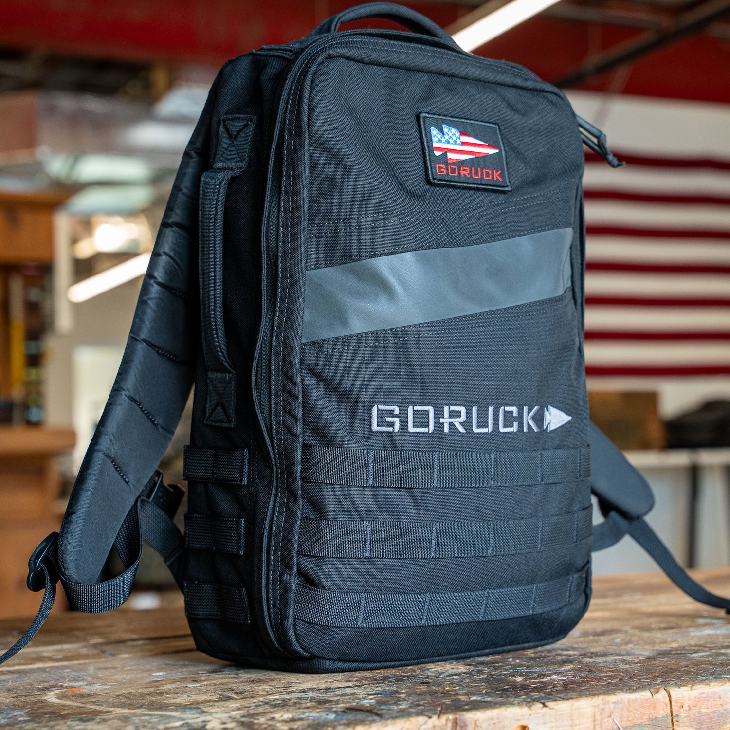 A black Rucker 5.0 rucksack with an American flag patch sits on a wooden table indoors, ready for your next adventure and designed with excellent lumbar support.