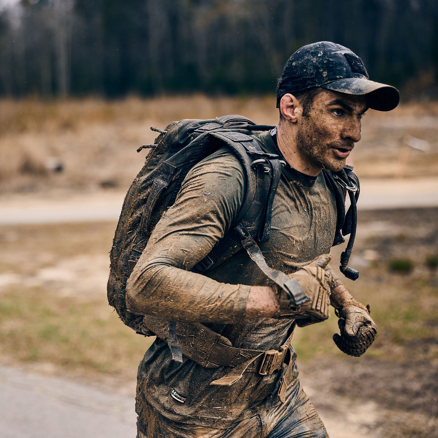 A man runs through a muddy, forested area with the Rucker 5.0 and a cap, enjoying the challenge of rucking and the enhanced lumbar support his gear offers.