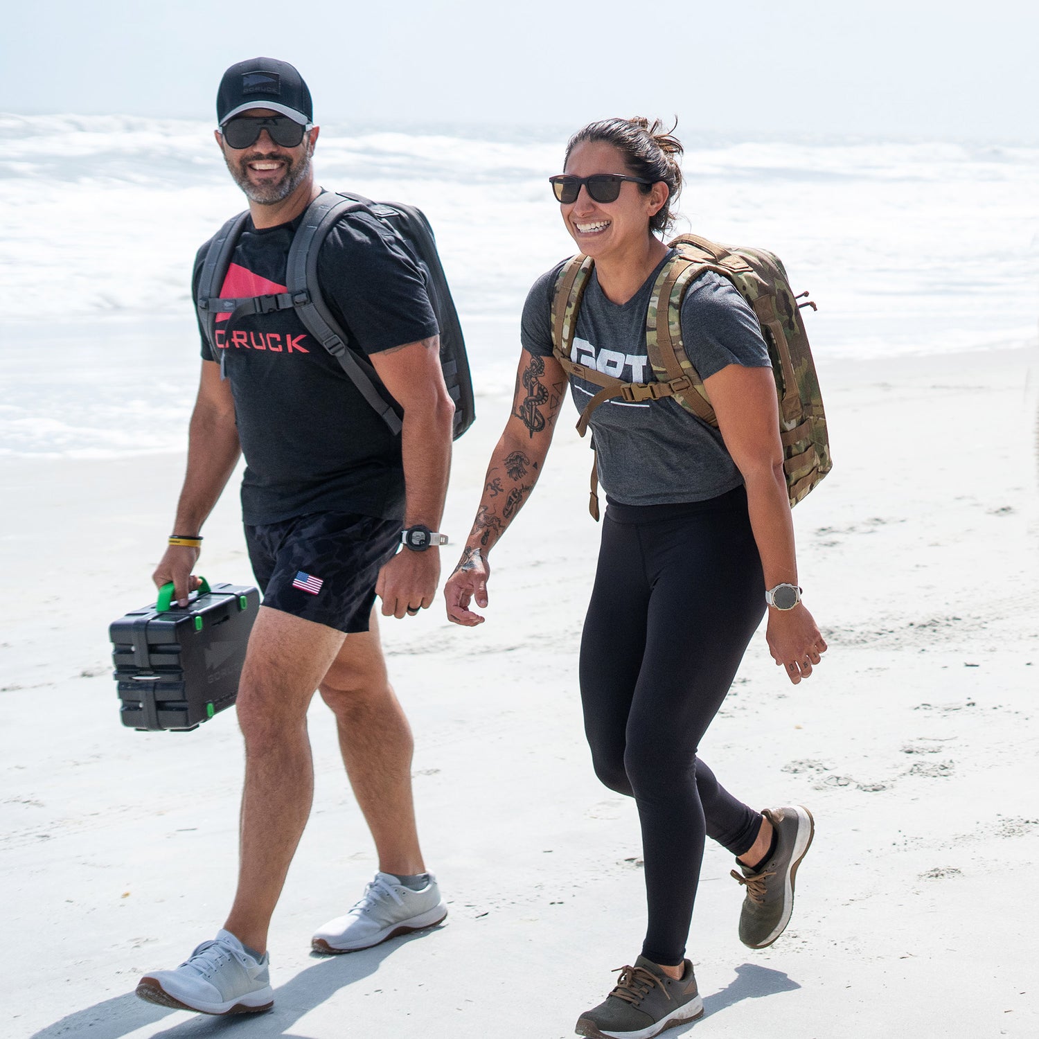 Two people with rucksacks walk and smile on a sunny beach, enjoying the ocean breeze and the superior lumbar support of the Rucker 5.0.