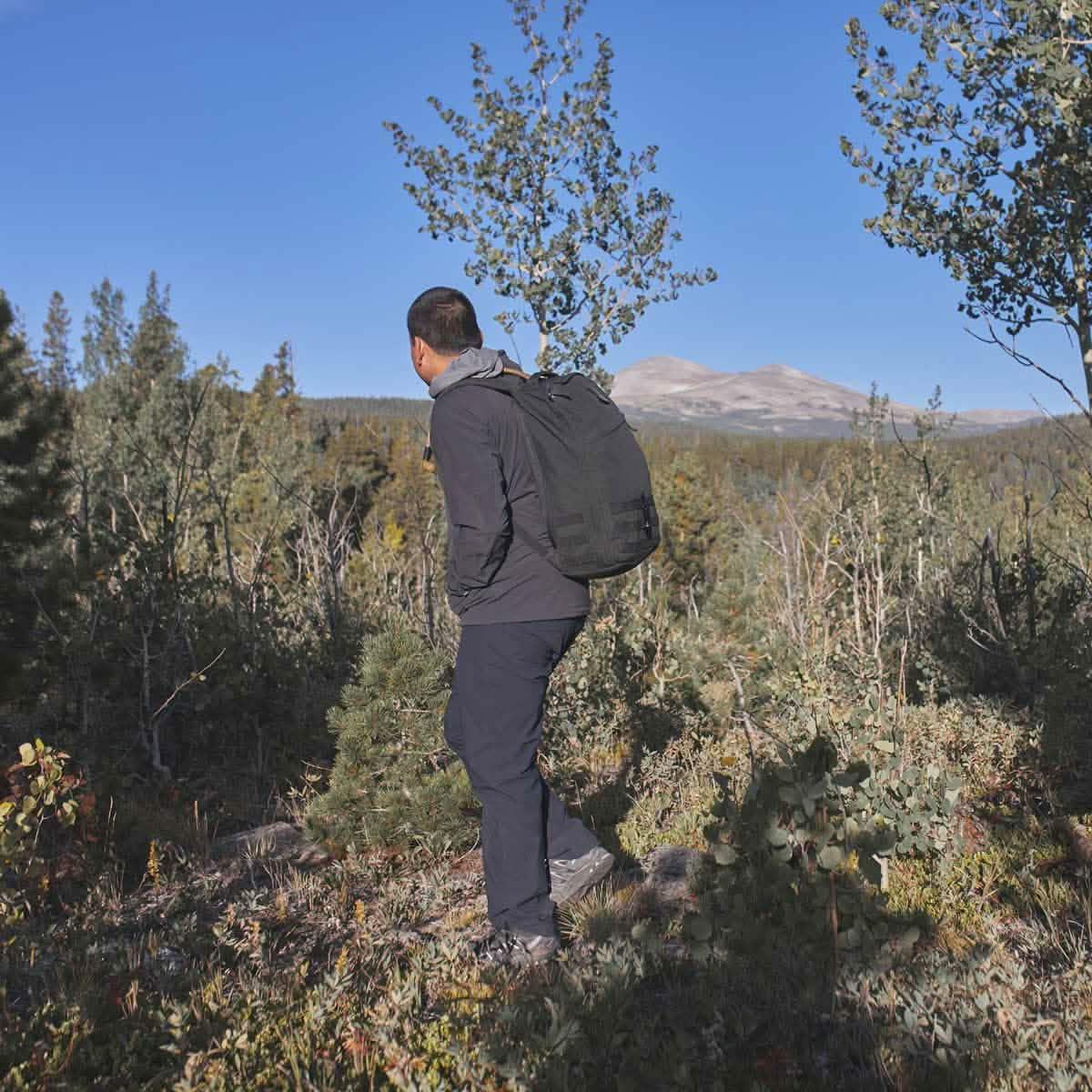 Man rucking in GORUCK gear and pants on a mountain trail surrounded by trees and blue sky.
