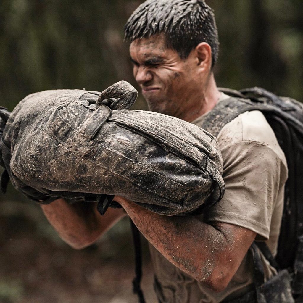A man covered in mud strains to lift a heavy, muddy sandbag outdoors.