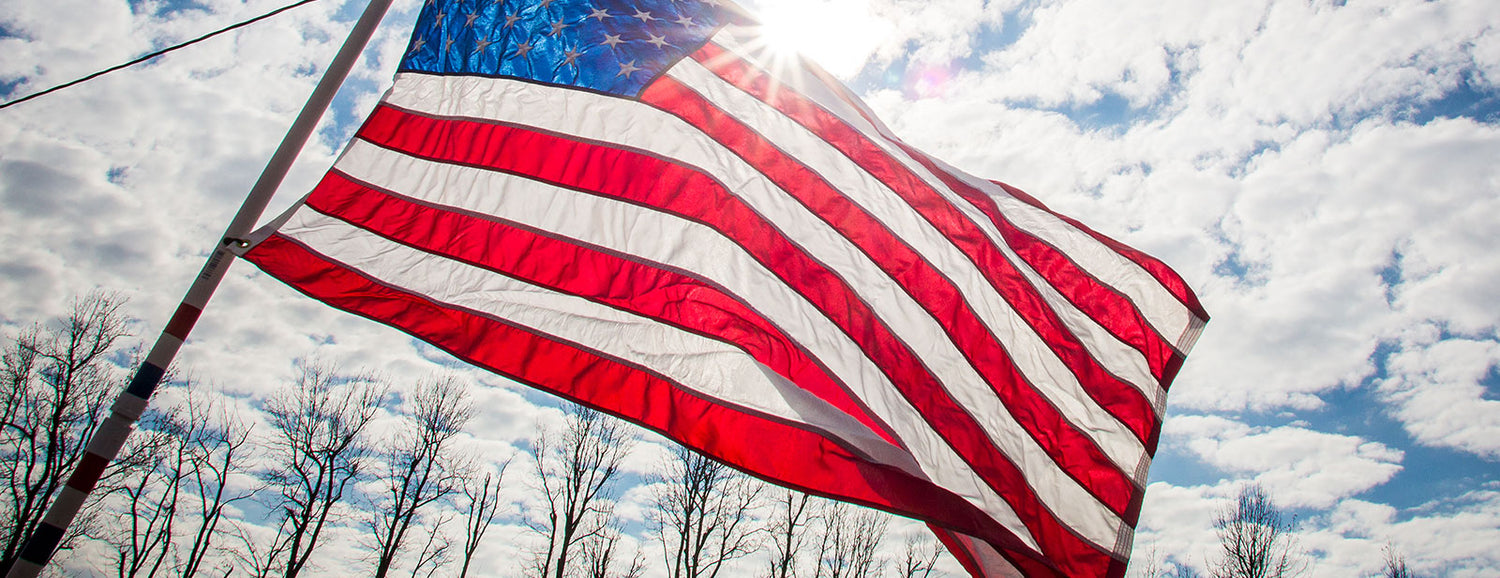 An American flag waves in the wind against a partly cloudy sky with sunlight shining through.