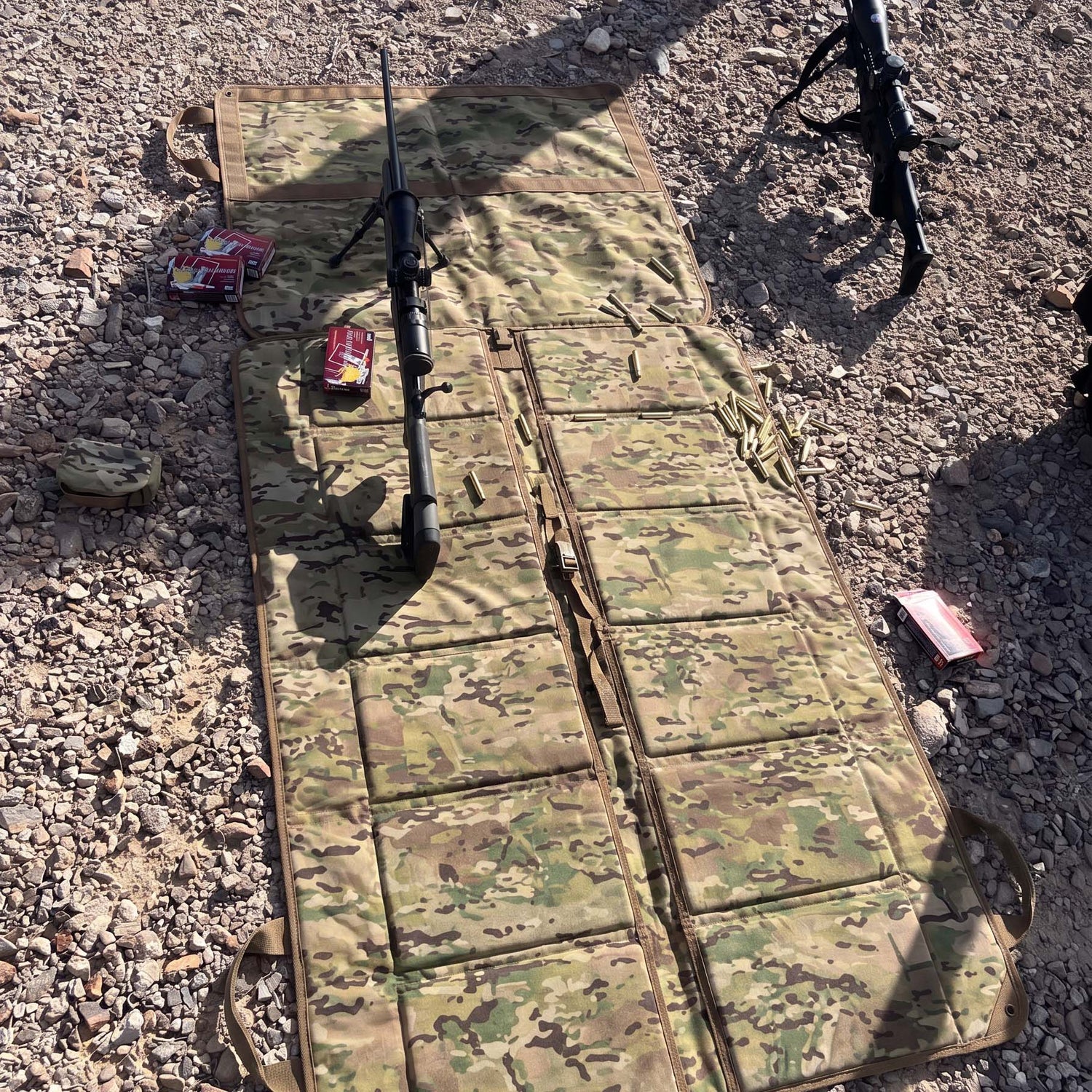 A Shooting Mat is spread outdoors on rocky ground, displaying a rifle and ammunition, with another rifle partially visible.
