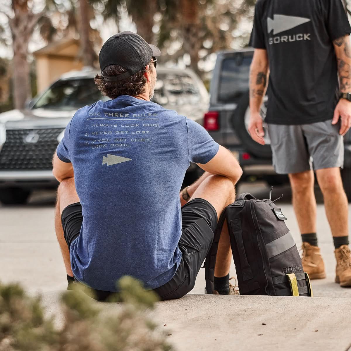Man wearing GORUCK Three Rules navy tee sitting outdoors with rucksack, another in logo shirt.