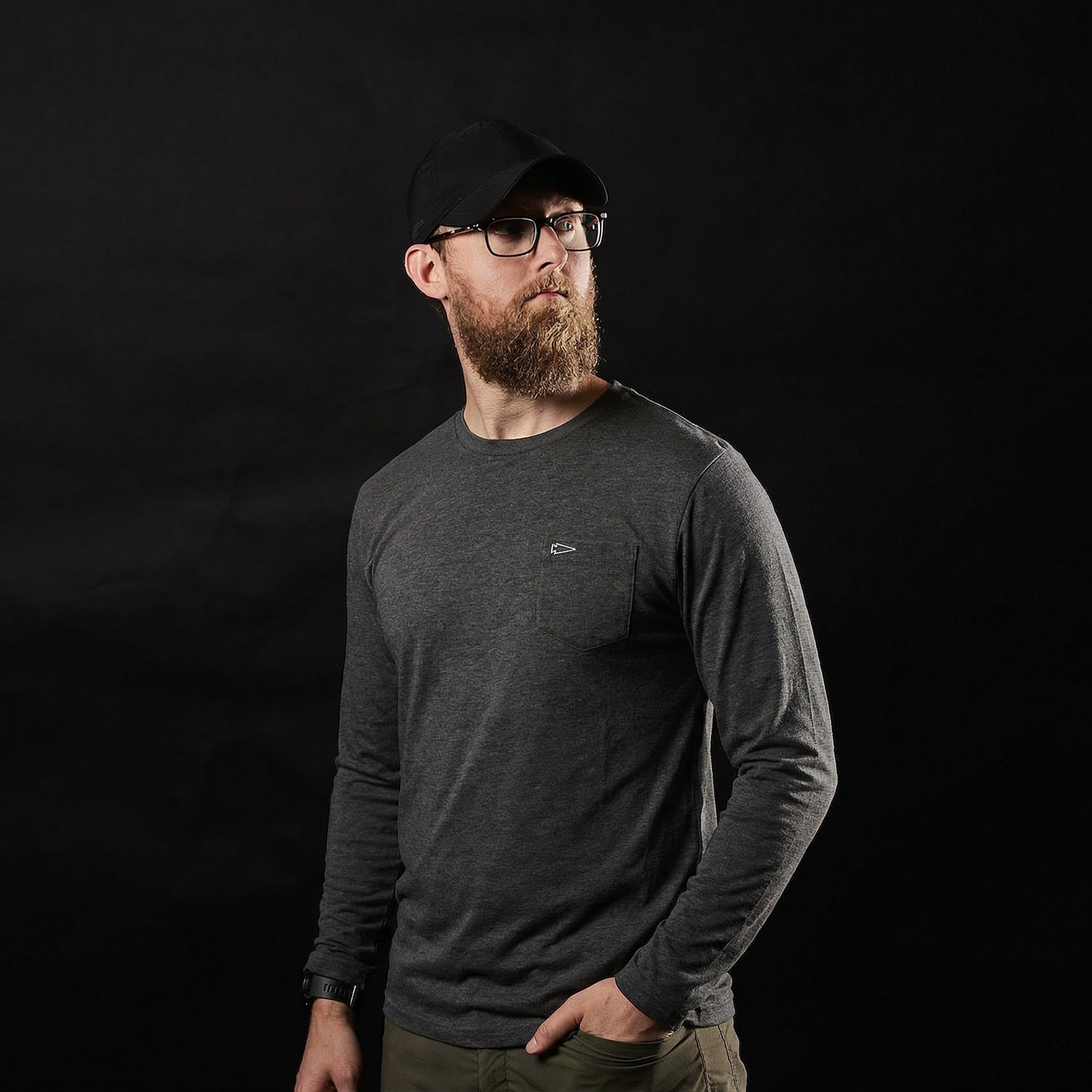 A man with a beard, wearing glasses, a black cap, and the Men's Long Sleeve Pocket Tee - Tri-BlendX in dark color stands against a black background.