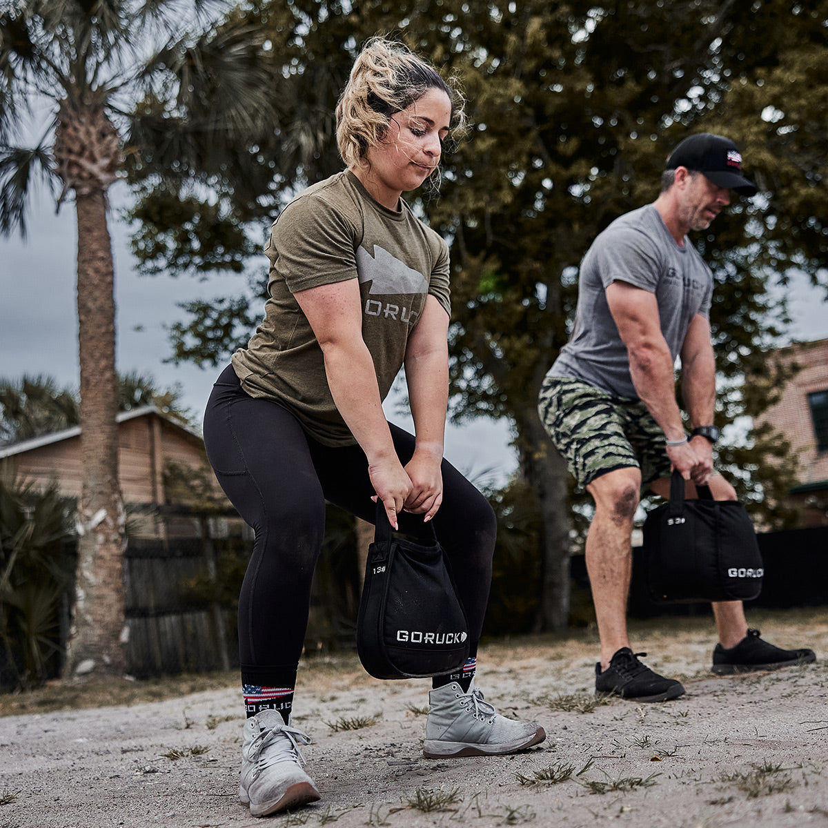 Two people outdoors lifting weighted bags, exercising on sand near trees and a house.