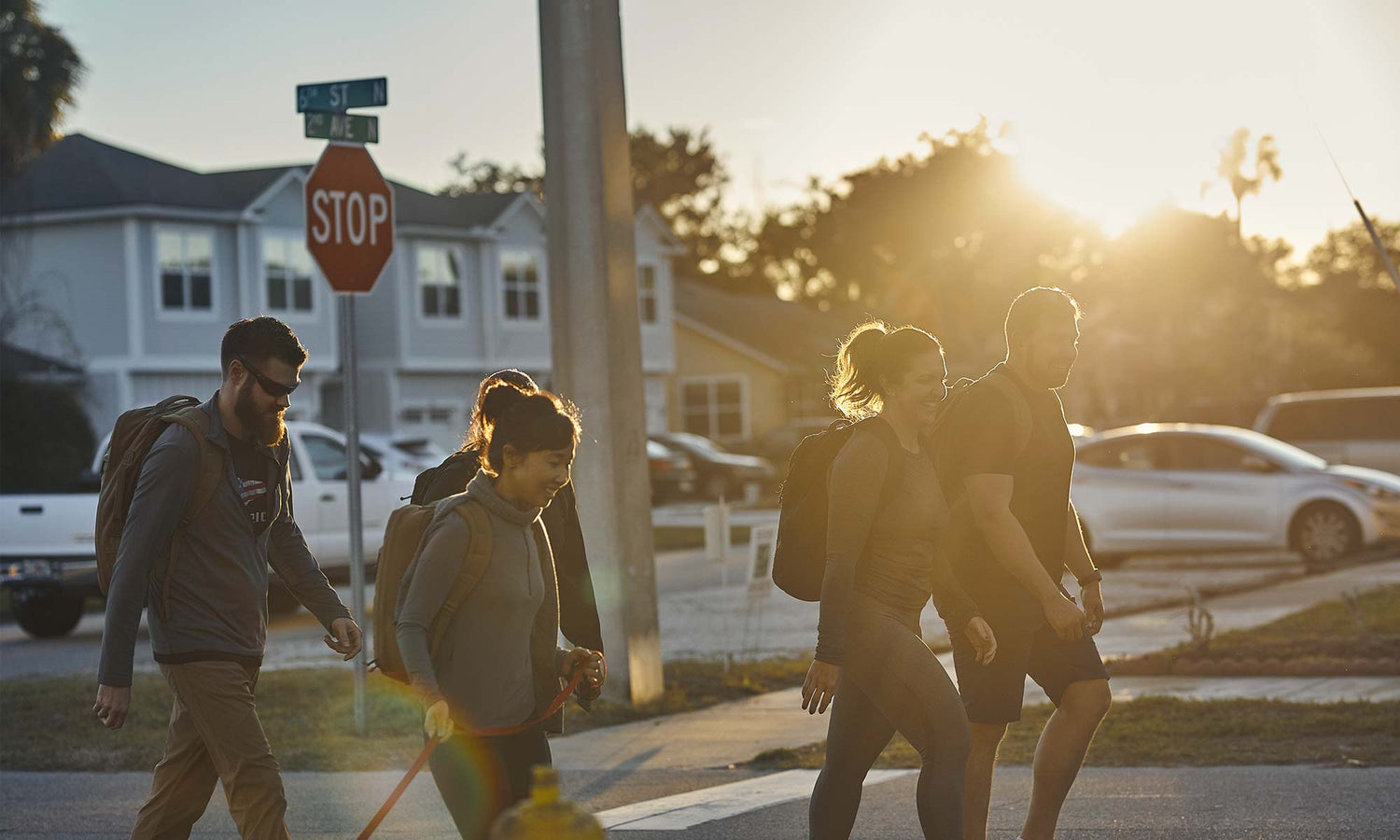 A group of people with backpacks walk together down a street at sunset near a stop sign.