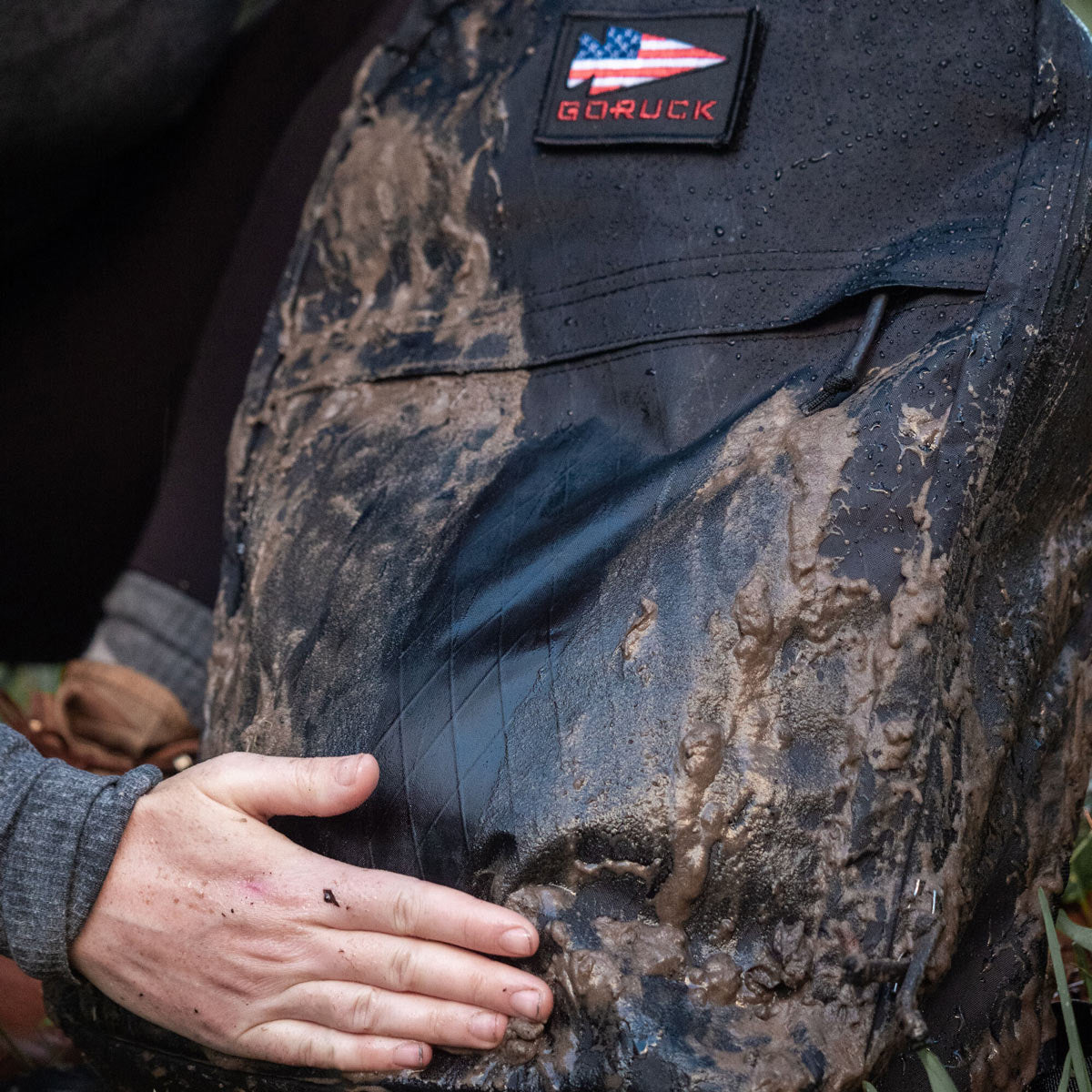 A muddy GORUCK backpack with a person’s hand wiping off dirt from the front.