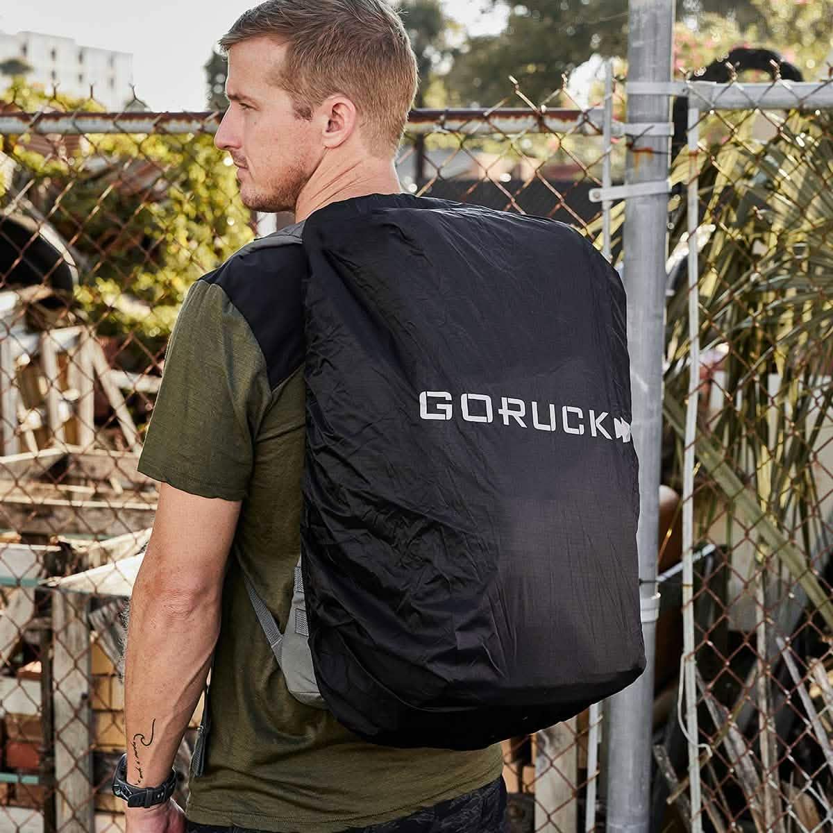 Man outdoors wearing black GORUCK backpack cover, rucking gear, chain link fence background