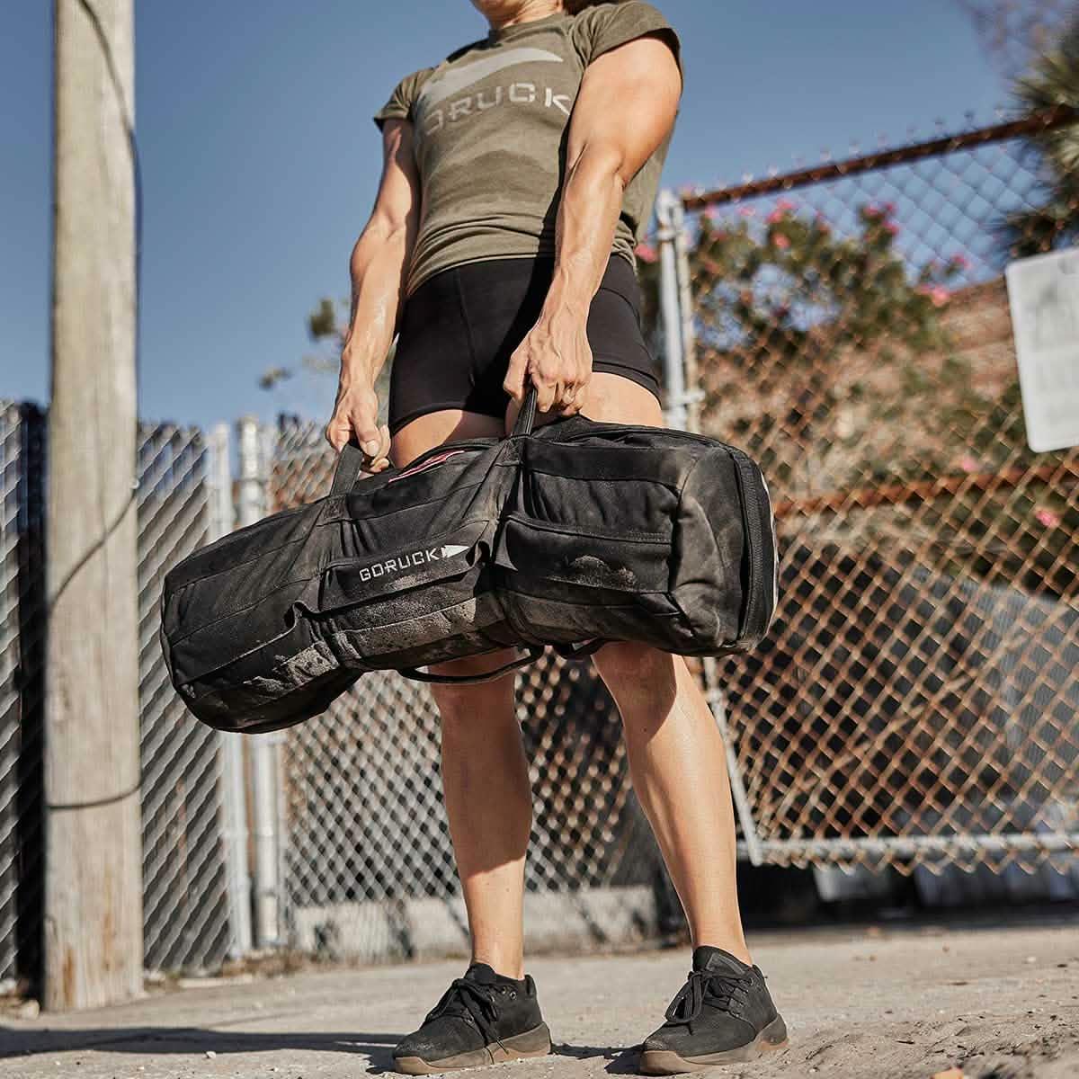 Athlete lifting GORUCK 80lb sandbag outdoors, showcasing durable rucking gear