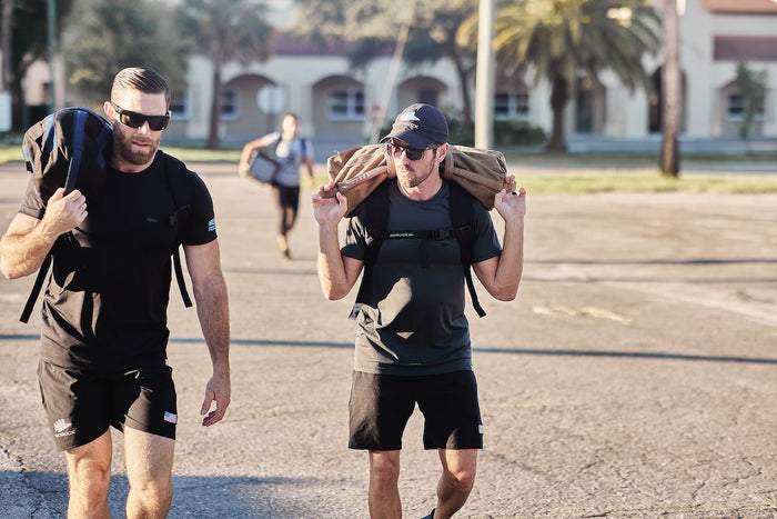 Two men rucking outdoors with GORUCK gear and sandbags, wearing athletic apparel