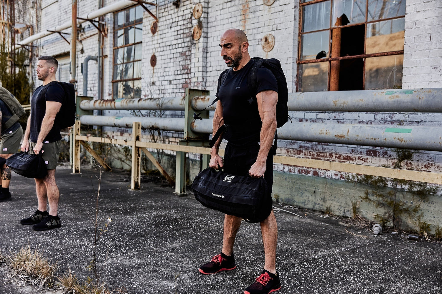 Men performing outdoor rucking workout with GORUCK backpacks and sandbags beside industrial building